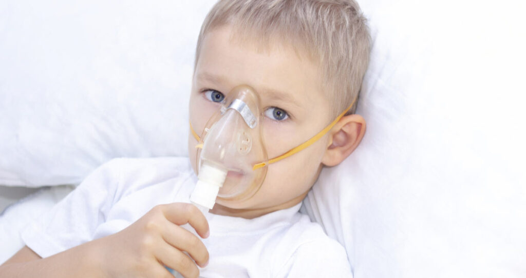 Niño con mascarilla de inhalador: problemas respiratorios por asma. Un niño con mascarilla de inhalador está acostado en la cama e inhala adrenalina. Concepto de atención sanitaria y niño enfermo; coronavirus, bronquitis, neumonía.
