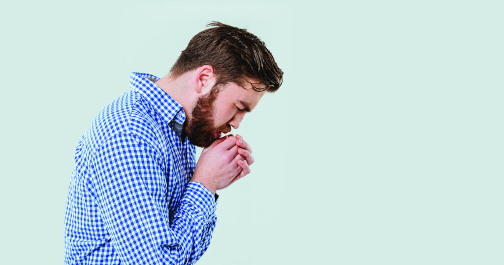 Un hombre con una camisa a cuadros azul y blanca parado de lado con la cabeza inclinada y las manos entrelazadas delante de la boca como si estuviera sumido en sus pensamientos o preocupación, sobre un fondo verde claro.