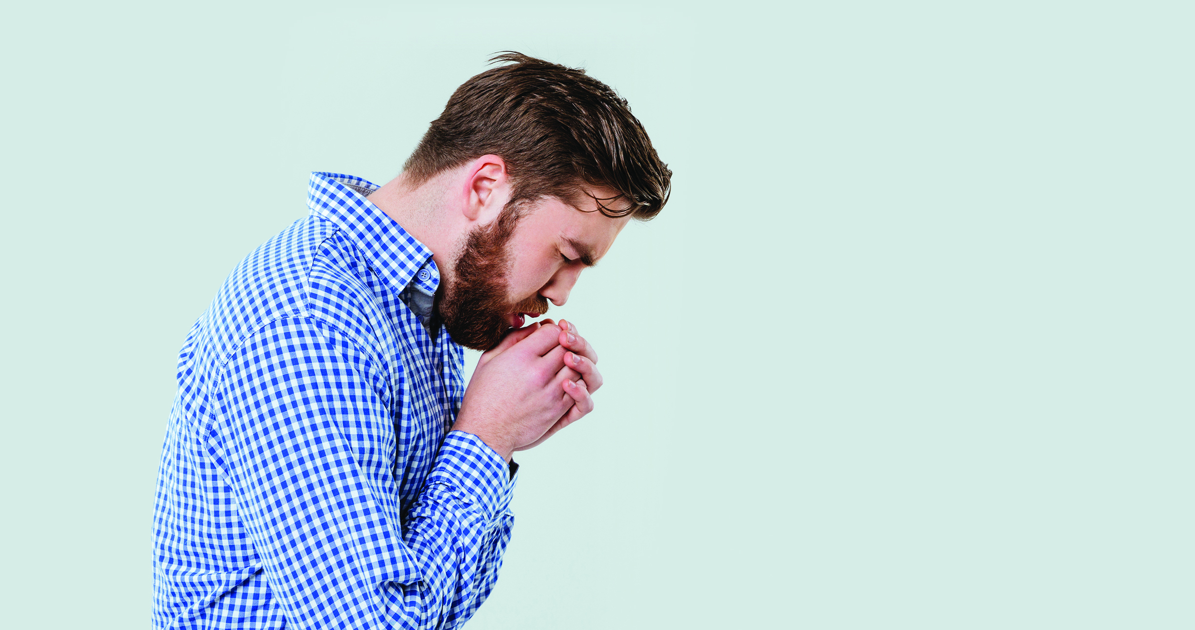 Un hombre con una camisa a cuadros azul y blanca parado de lado con la cabeza inclinada y las manos entrelazadas delante de la boca como si estuviera sumido en sus pensamientos o preocupación, sobre un fondo verde claro.