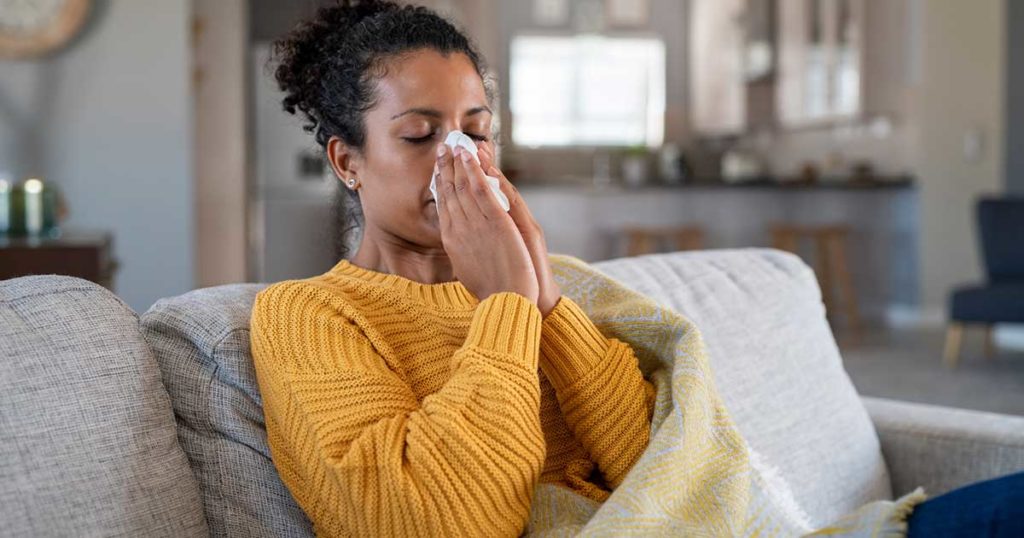 Mujer sentada en un sillón sonándose la nariz con ambas manos y un pañuelo. Sufre de sinusitis.