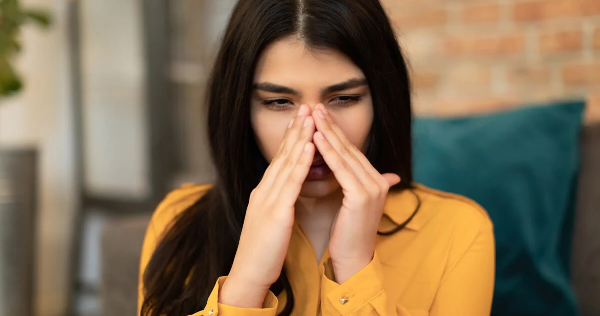 Una mujer joven con una blusa amarilla parece angustiada con los dedos en las sienes, posiblemente experimentando dolor de cabeza o presión en los senos nasales.