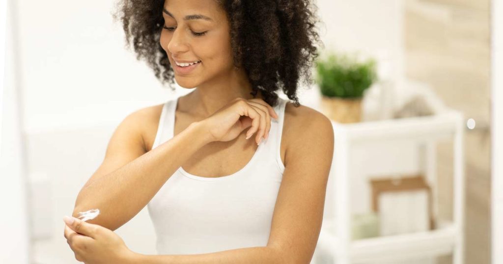 Joven mujer negra, sonriente, con camiseta sin mangas, aplicándose crema para el eczema en el codo