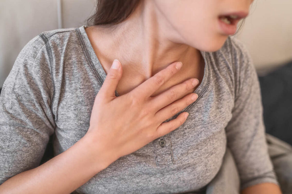 Una mujer se toca el pecho con la boca abierta. Se nota esfuerzo en el cuello porque tiene dificultad para respirar a causa de la aspergilosis broncopulmonar alérgica (ABPA).