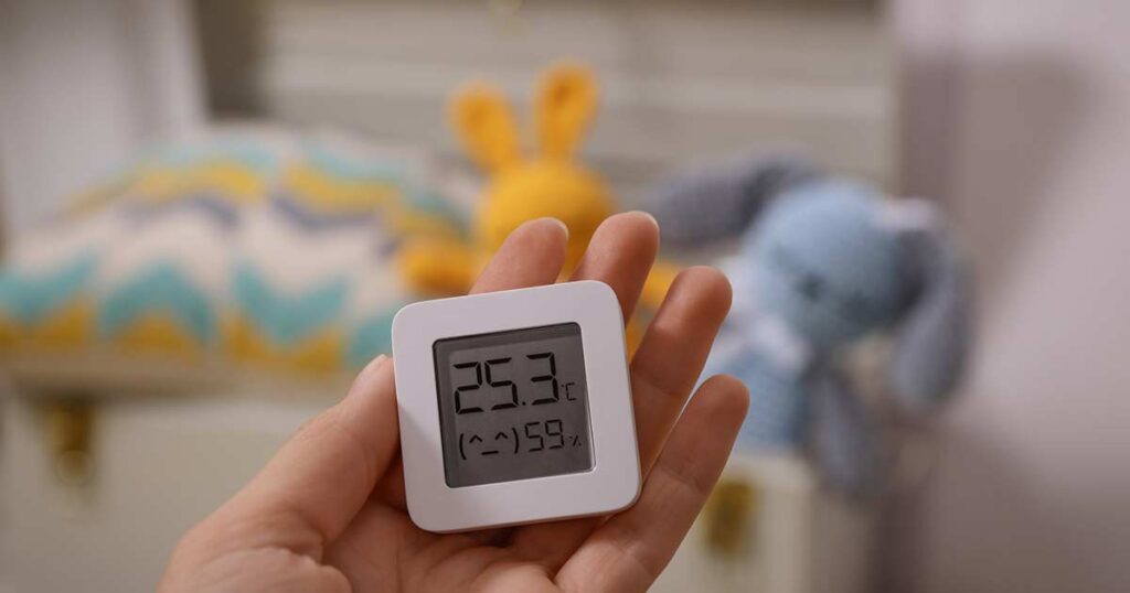 Una mano sostiene un termómetro digital que indica 25,3 °C y una humedad del 59 %. En el fondo borroso se ven juguetes de peluche en tonos amarillos, azules y grises.