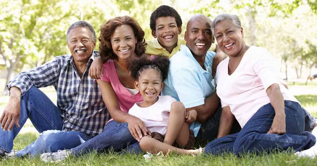 Seis integrantes de una familia multigeneracional (tres adultos y tres niños) están sentados juntos en el césped de un parque. Están sonriendo y visten informalmente. Al fondo se ven frondosos árboles verdes, que sugieren un día soleado.