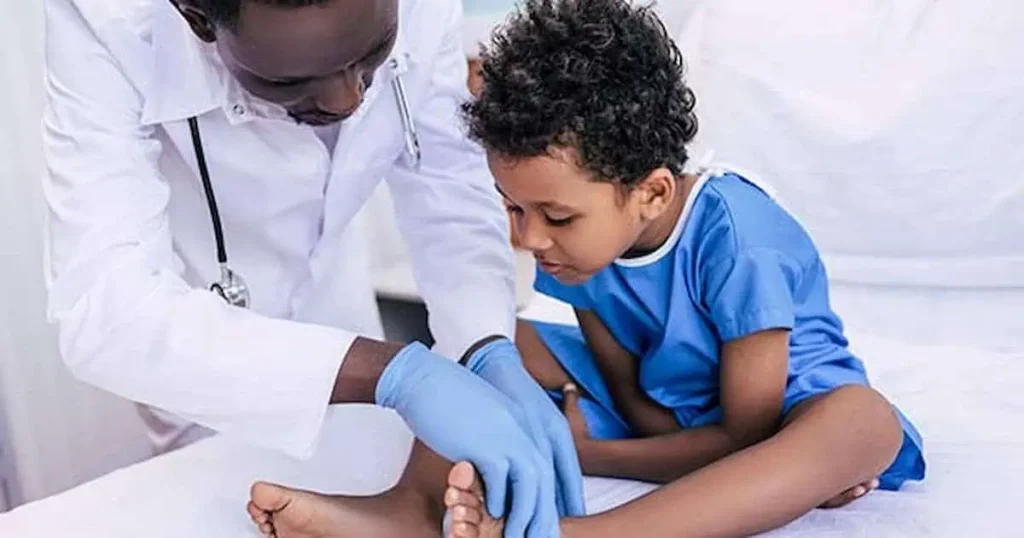Un médico con guantes examina el pie de un niño pequeño sentado en una camilla médica. El niño lleva una bata azul y parece concentrado en el examen. El entorno parece ser una clínica.