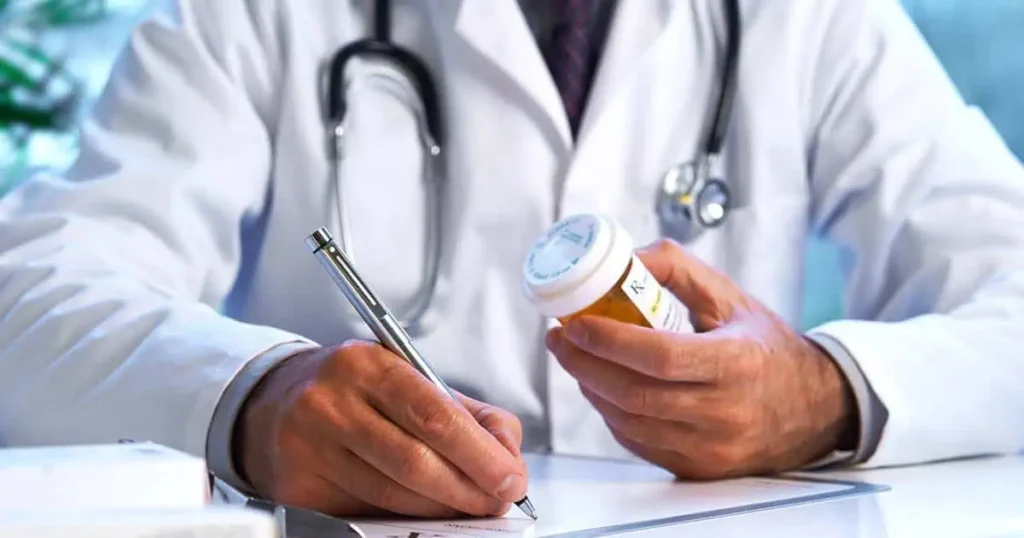 Un médico con bata blanca y estetoscopio escribe una receta mientras sostiene un frasco de pastillas en un escritorio.