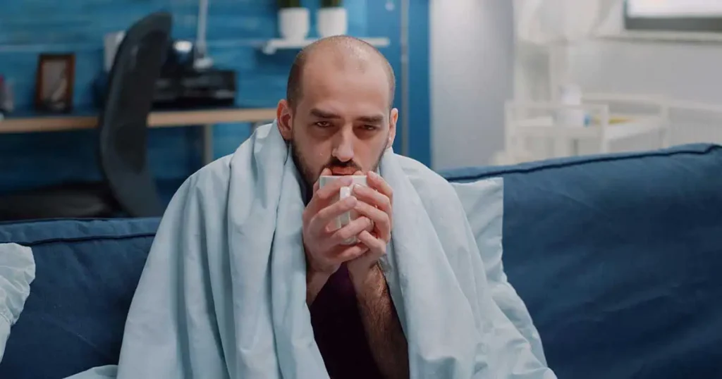 Un hombre con una manta envuelta alrededor de los hombros está sentado en un sofá, sosteniendo una taza con ambas manos. Parece cansado o indispuesto, con una expresión pálida, en un ambiente hogareño y acogedor.