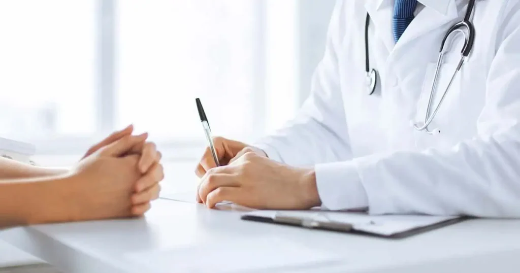 Un médico con bata blanca y estetoscopio escribe en un portapapeles mientras está sentado en un escritorio frente a un paciente con las manos cruzadas.
