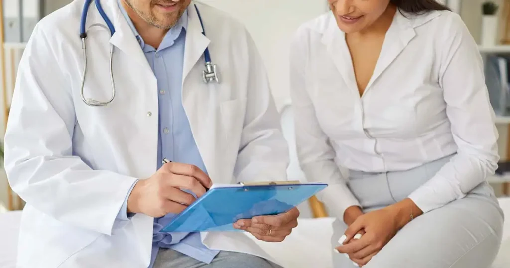 Un médico con bata blanca revisa notas en un portapapeles mientras está sentado junto a un paciente, ambos vestidos con camisas blancas, en un consultorio médico.