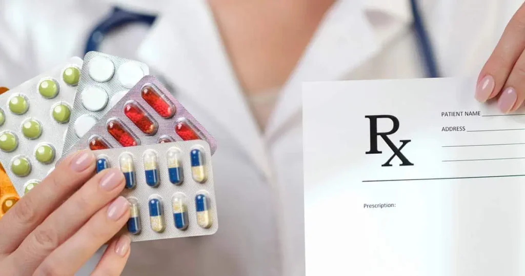 Una persona con bata blanca sostiene en una mano blísters con diferentes pastillas y en la otra un formulario de receta médica en blanco, sugiriendo un contexto médico o farmacéutico.