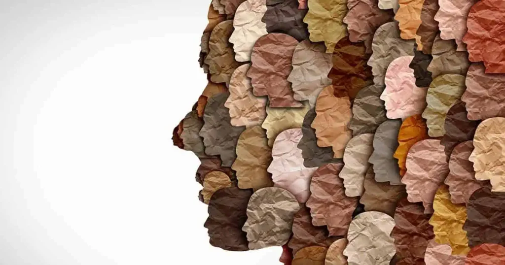 Siluetas de cabezas humanas en varios tonos de piel forman un gran perfil sobre un fondo blanco, simbolizando la diversidad, la unidad y la inclusión. Las cabezas tienen una textura que parece papel arrugado.
