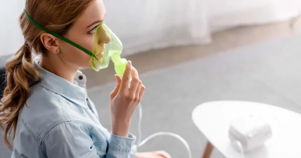 Una mujer de cabello rubio, usando una mascarilla de oxígeno verde, está sentada en el interior de una habitación. Sostiene en la mano una boquilla de nebulizador conectada a una máquina sobre una mesa blanca cercana. Mira hacia un lado, con una expresión de calma y concentración.