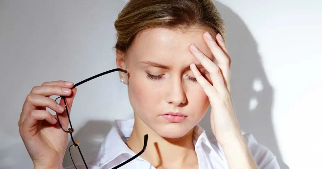 Una mujer con los ojos cerrados sostiene sus gafas en una mano y se toca la frente con la otra, aparentando estar estresada o con dolor de cabeza.