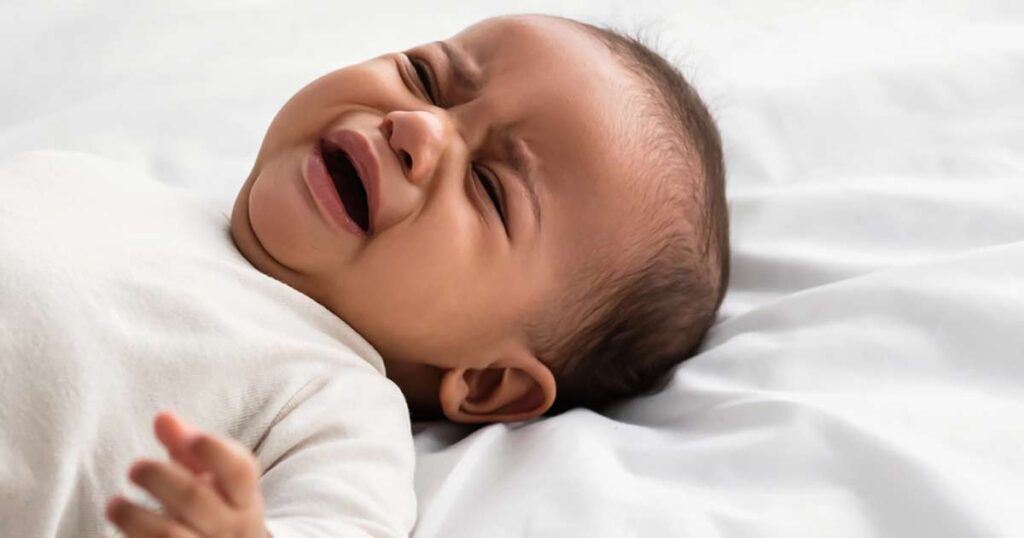 Un bebé acostado en una cama blanca, vestido con ropa de color claro, con los ojos cerrados y la boca abierta, parece estar llorando o molesto, lo que podría ser un signo temprano de anafilaxia infantil.