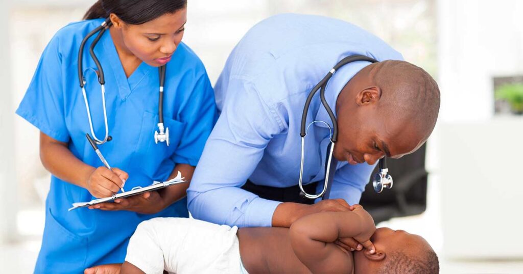 Un médico examina a un bebé acostado en una camilla mientras una enfermera con uniforme azul está cerca, tomando notas en un portapapeles. Ambos adultos llevan estetoscopios.