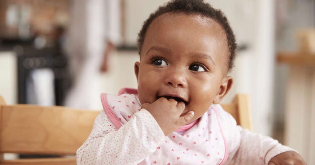 Un bebé vestido con ropa rosa y blanca sentado en una silla alta, sonriendo y mordiéndose los dedos: un momento adorable que nos recuerda que debemos estar atentos a los signos de anafilaxia infantil. El fondo suavemente borroso mantiene el énfasis en el bebé.