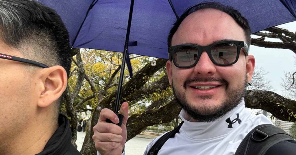 Un hombre con gafas de sol y una chaqueta blanca de Under Armour sonríe a la cámara mientras sostiene un paraguas azul. Otra persona está de pie junto a él, parcialmente fuera de plano. Al fondo se ven árboles sin hojas.