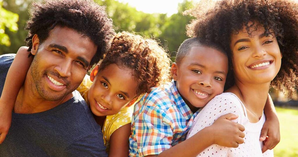 Una familia feliz de cuatro posa al aire libre, sonriendo a la cámara. Dos adultos y dos niños están muy juntos bajo la brillante luz del sol, con árboles verdes al fondo.