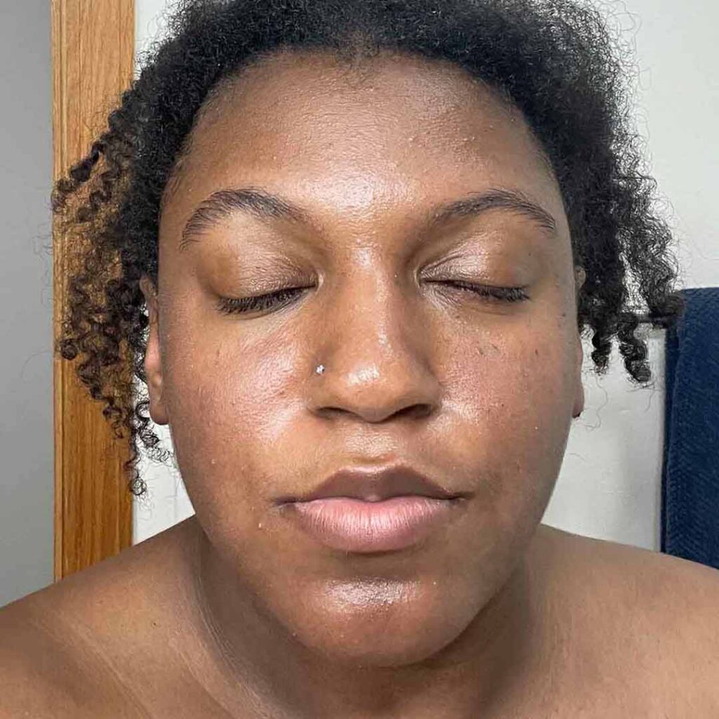 Una persona con los ojos cerrados y cabello natural está de pie en el interior, mirando a la cámara. Su piel luce tersa y luminosa, y tiene una expresión relajada y tranquila.