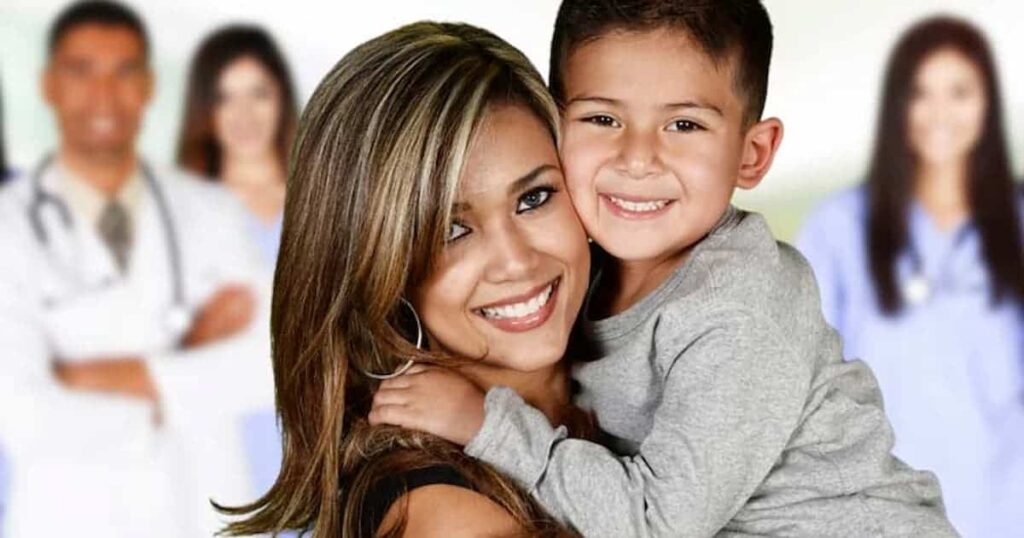 Una mujer sonriente sostiene a un niño pequeño; ambos miran a la cámara. Al fondo, ligeramente desenfocado, se distinguen varias personas con uniformes médicos, lo que sugiere un entorno sanitario.