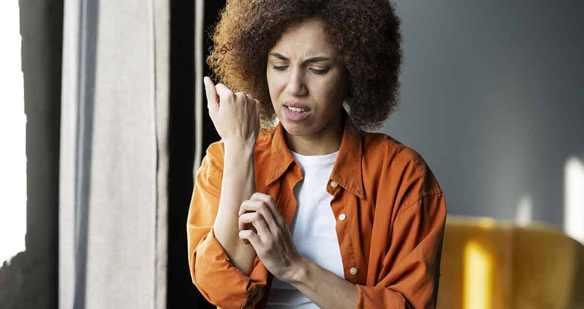 Una mujer con el pelo rizado y una camisa naranja se mira el brazo con incomodidad y se rasca el antebrazo, posiblemente por picazón o irritación, mientras está de pie en el interior cerca de una ventana.