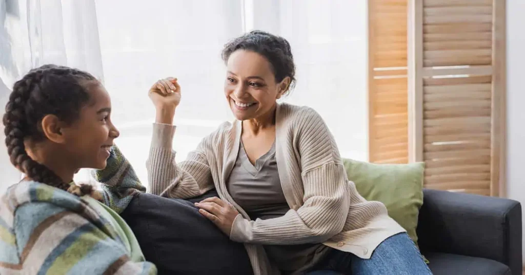 Una mujer con un cárdigan beige está sentada en un sofá, sonriendo y hablando con una niña de trenzas envuelta en una cobija a rayas. Están en una habitación luminosa con rayos de sol entrando por la ventana.