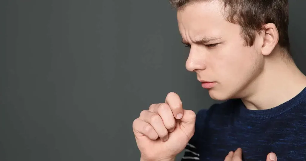 Un joven de piel clara y cabello corto aparece de perfil contra un fondo gris oscuro. Lleva un suéter azul marino y tose con el puño cerrado, luciendo incómodo o pensativo.