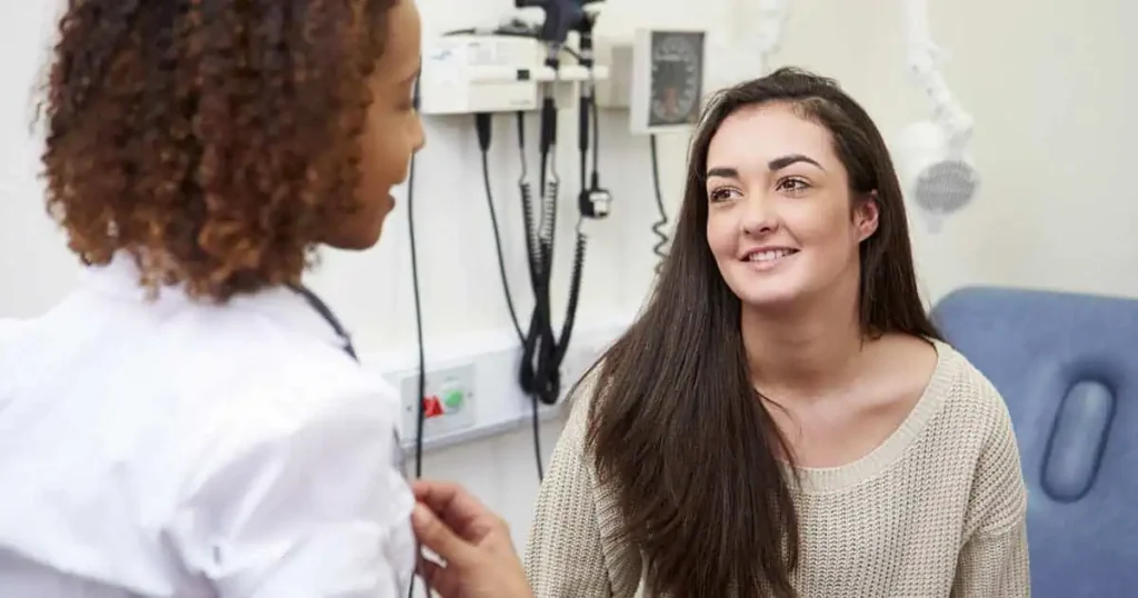 Una médica de cabello rizado habla con una paciente de cabello largo y oscuro en un consultorio. La paciente, que usa un suéter claro, está sentada y sonríe. Se ve equipo médico de fondo.