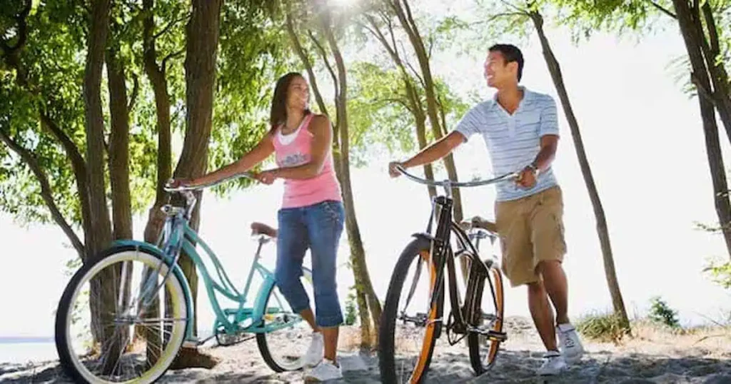 Una mujer y un hombre caminando con bicicletas por una zona boscosa e iluminada por el sol. La mujer, con una camiseta rosa, y el hombre, con una camisa a rayas, se sonríen el uno al otro. La luz del sol se filtra a través de los árboles, proyectando sombras moteadas en el suelo.