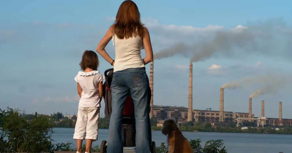 Una mujer está parada con un niño y un cochecito, acompañada por un perro, mirando a través de un cuerpo de agua. Al fondo se ven varias chimeneas industriales humeantes bajo un cielo despejado.