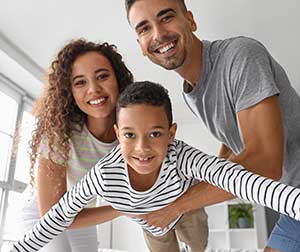 Una mujer y un hombre sonrientes sostienen a un niño alegre que tiene los brazos extendidos como si estuviera volando; los tres miran a la cámara en una sala de estar luminosa y moderna.