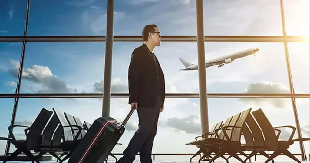 Un hombre con traje camina por una terminal de aeropuerto, llevando una maleta. Se ven filas de asientos vacíos y, a través de unos grandes ventanales que enmarcan un cielo brillante, se ve un avión despegando.