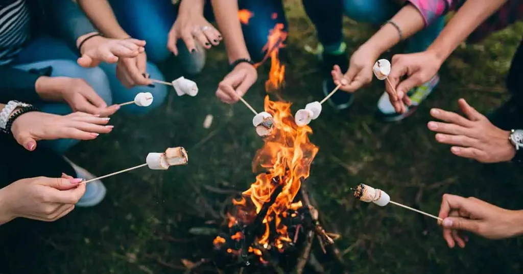 Un grupo de personas sentadas alrededor de una fogata en un campamento de verano, asando malvaviscos en palos sobre las llamas. Solo se ven sus manos y palos, con kits de epinefrina cerca, mientras el fuego arde brillantemente en el centro.