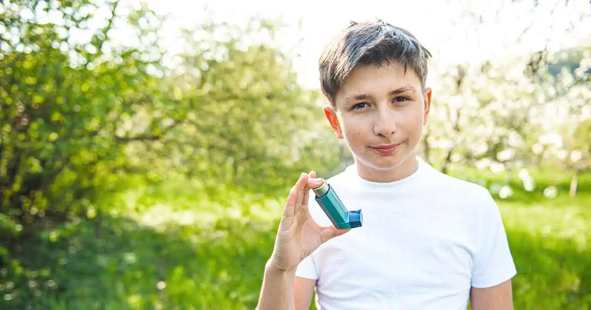 Un niño parado al aire libre en un entorno verde y soleado sostiene un inhalador para el asma, y tiene puesta una camisa blanca.