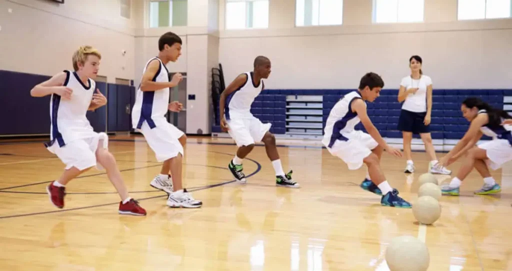 Un grupo de cuatro jóvenes atletas con uniformes azules y blancos participan en un ejercicio de baloncesto en un gimnasio, con un entrenador que observa desde el fondo. Dos jugadores se lanzan hacia las pelotas de baloncesto que están en el suelo.