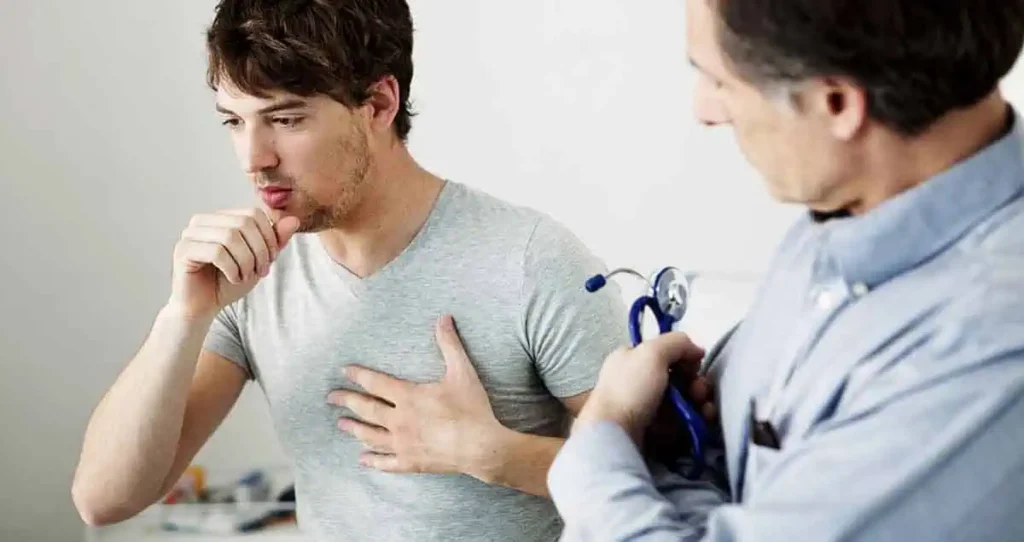 Un joven que lleva una camiseta gris tose y se toca el pecho. Se encuentra en una consulta con un médico, que lo observa y sostiene un estetoscopio.