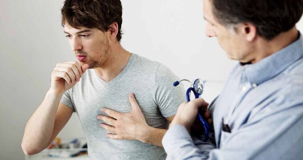 Un joven que lleva una camiseta gris tose y se toca el pecho. Se encuentra en una consulta con un médico, que lo observa y sostiene un estetoscopio.