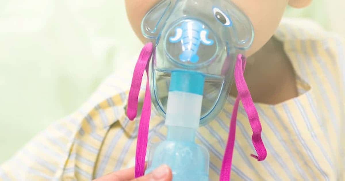 Un niño pequeño con una camiseta de rayas utiliza una mascarilla de nebulización azul con diseño de elefante y correas rosas. El niño sostiene el nebulizador con seguridad, lo que sugiere que está acostumbrado a usarlo.