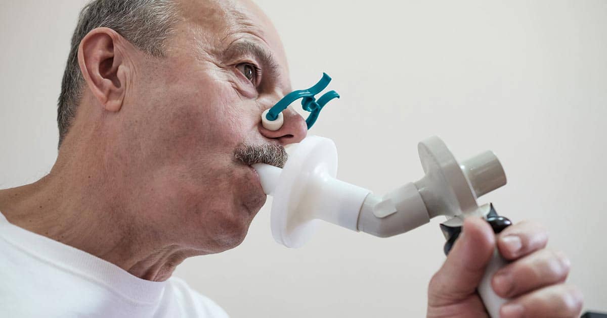 Una persona se somete a una prueba de función pulmonar para el asma utilizando un espirómetro con boquilla y pinza nasal. Se le ve concentrada en la actividad, lo que sugiere un entorno de evaluación médica o sanitaria.