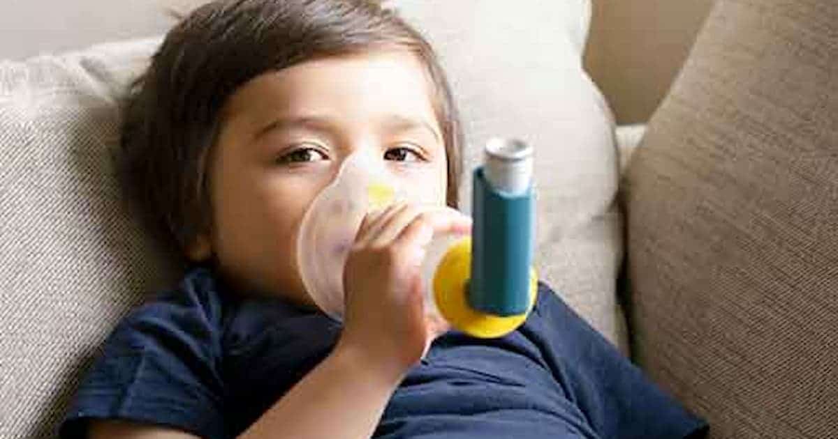 Un niño está tumbado en un sofá usando un inhalador con mascarilla. Tiene el pelo corto y castaño, y lleva una camiseta azul oscuro. El inhalador es azul con un accesorio amarillo, y la escena parece desarrollarse en un interior con muebles de tonos neutros.
