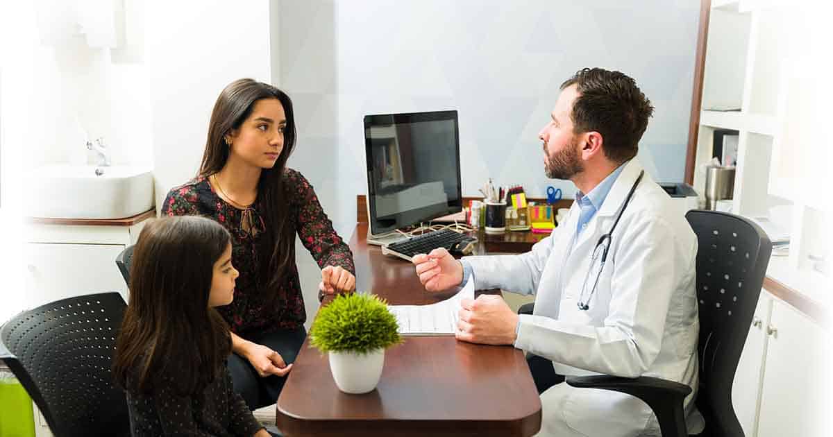 Un médico con bata blanca está sentado a un escritorio hablando con una mujer y una niña. La mujer está sentada en una silla y la niña está sentada junto a ella. Al fondo se ve un despacho con una computadora y una planta.